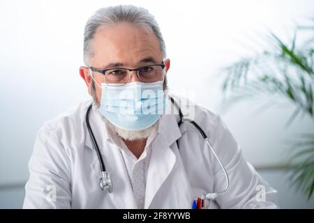 Portrait d'un médecin barbu en manteau blanc et masque facial regardant l'appareil photo dans son bureau. Banque D'Images