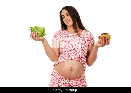 Femme enceinte qui choisit entre une laitue et un hamburger, isolée sur fond blanc