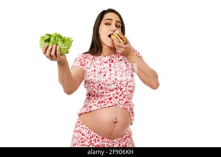 Femme enceinte qui choisit entre une laitue et un hamburger, isolée sur fond blanc