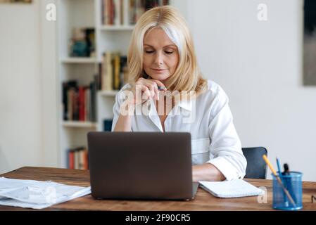 Confiante belle femme blonde caucasienne senior, responsable ou femme d'affaires, dans chemise blanc élégant, utilise le lapto, travaillant à distance sur le projet d'affaires, pensant à la stratégie, regarde l'écran d'ordinateur portable Banque D'Images