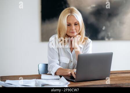 Blond de race blanche mature, femme d'affaires, Manager ou freelance, portant un maillot blanc, travaillant à la maison avec un ordinateur portable au-dessus d'une idée d'entreprise, développer une stratégie, regarde l'écran d'ordinateur portable Banque D'Images