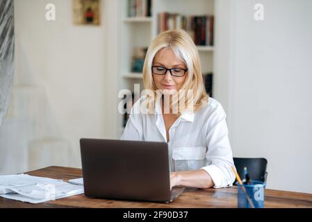 Concentrée influente belle femme blonde caucasienne mûre, portant des lunettes et chemise blanche, pdg, femme d'affaires ou courtier travaillant sur ordinateur portable, faire rapport, développer la stratégie, taper sur le clavier Banque D'Images