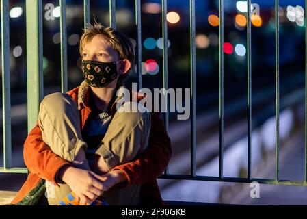 Adolescent avec masque regardant loin assis contre le rail au-dessus du train pistes Banque D'Images