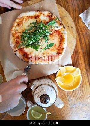 Mains d'un homme coupant pizza, thé, citron sur une table en bois dans un café Banque D'Images