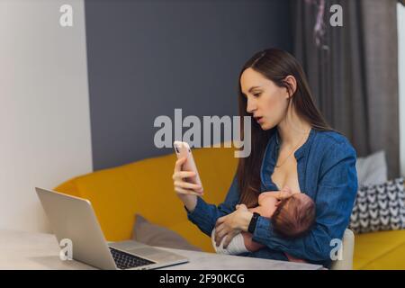 Belle jeune mère travaillant avec un ordinateur portable et allaitant, tenant et allaitant son nouveau-né à la maison. Maman - femme d'affaires nourrissant newb Banque D'Images