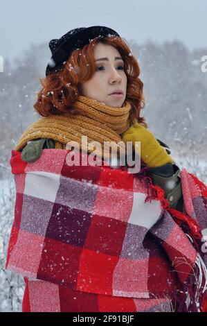 Joyeuse fille aux cheveux rouges réchauffe ses mains enveloppées dans un chaud carreaux rouges dans un champ lors d'une chute de neige Banque D'Images