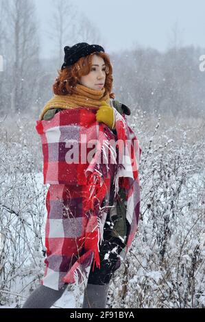 Joyeuse fille aux cheveux rouges réchauffe ses mains enveloppées dans un chaud carreaux rouges dans un champ lors d'une chute de neige Banque D'Images