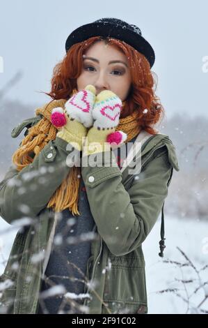 Une jeune fille gaie aux cheveux rouges réchauffe ses mains dans des moufles tricotées chaudes dans le champ pendant une chute de neige Banque D'Images