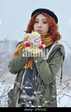 Une jeune fille gaie aux cheveux rouges réchauffe ses mains dans des moufles tricotées chaudes dans le champ pendant une chute de neige Banque D'Images