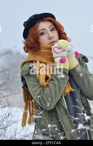 Une jeune fille gaie aux cheveux rouges réchauffe ses mains dans des moufles tricotées chaudes dans le champ pendant une chute de neige Banque D'Images