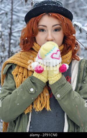 Une jeune fille gaie aux cheveux rouges réchauffe ses mains dans des moufles tricotées chaudes dans le champ pendant une chute de neige Banque D'Images