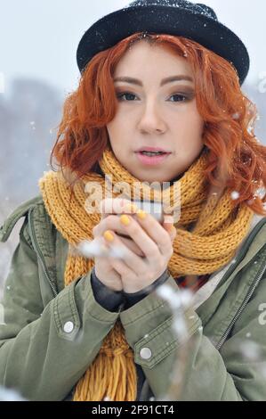 Une jeune fille joyeuse aux cheveux rouges se réchauffe les mains et boit du thé chaud d'un thermos mug dans le champ lors d'une chute de neige Banque D'Images