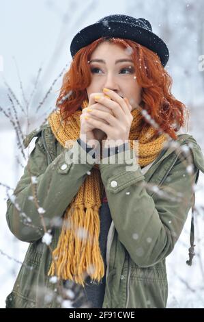 Une jeune fille joyeuse aux cheveux rouges se réchauffe les mains et boit du thé chaud d'un thermos mug dans le champ lors d'une chute de neige Banque D'Images