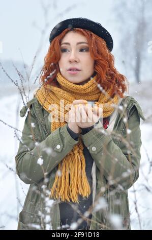 Une jeune fille joyeuse aux cheveux rouges se réchauffe les mains et boit du thé chaud d'un thermos mug dans le champ lors d'une chute de neige Banque D'Images