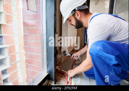 Vue latérale d'une combinaison de travail de jeune homme barbu qui installe un radiateur de chauffage dans la pièce. Homme travaillant dans un casque de sécurité à l'aide d'une clé dans l'appartement. Concept de l'installation du radiateur et des travaux de plomberie. Banque D'Images