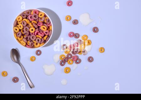 Des anneaux de maïs colorés dans un bol sur fond coloré. Les cornflakes sont dispersés sur la table en gouttes de lait. Vue de dessus. Banque D'Images