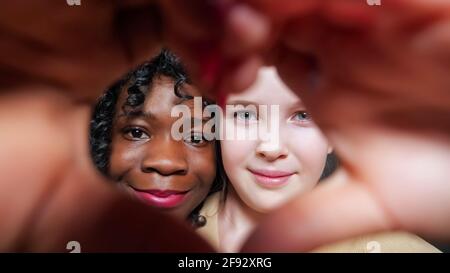 Belle-mère noire aux cheveux bouclés et blonde adopté école fille spectacle le cœur signe avec les mains et regarder dans les yeux avec joie sourit en gros plan Banque D'Images