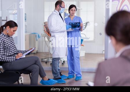 Dentiste dans la salle d'attente en stomatologie tenant une radiographie du patient expliquant le diagnostic à l'infirmière dans un couloir encombré avec la forme de remplissage du patient. Personnel de Dentistiry à la réception. Banque D'Images