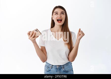 Femme soulagée et heureuse criant oui et recherchant satisfaite, tenant le smartphone, gagnant en ligne sur le téléphone mobile, atteindre l'objectif d'application, gagner des résultats Banque D'Images