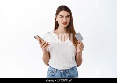 Elle déteste payer avec la carte de crédit pour les commandes. La jeune femme confuse tient carte de crédit et téléphone mobile, frowning et regardant déçu à l'appareil photo Banque D'Images