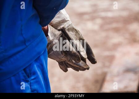 Gros plan de gants de travail sales sur fond de sol jaune désertique flou. Ouvrier de l'huile en vêtements de travail bleus. Banque D'Images