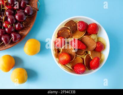 Mini crêpes aux fraises dans un bol, à côté des abricots et des cerises sur fond bleu avec un endroit pour le texte. Vue de dessus. Banque D'Images