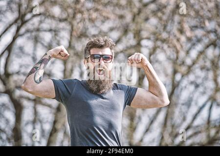 L'homme barbu montre des muscles, des biceps le jour ensoleillé. Concept de masculinité. L'homme avec une barbe longue a l'air énergique et gai. Homme avec barbe et moustache sur Banque D'Images