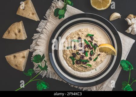 Cuisine arabe : Baba Ganoush avec olives noires et persil haché et pain plat sur fond noir, vue du dessus Banque D'Images