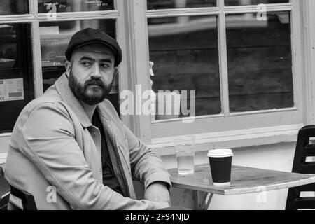 À York, un homme barbu dans un chapeau, se détendit à une table extérieure solitaire avec une boisson d'eau et une tasse de café, North Yorkshire, Royaume-Uni. Banque D'Images