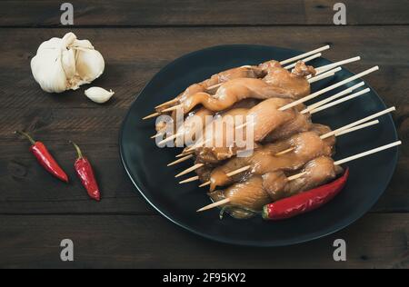 Assiette avec brochettes de poulet crues, ail et piment rouge sur bois sombre. Banque D'Images