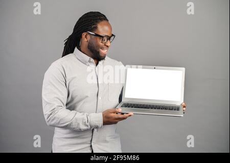 Homme noir de race mixte avec des dreadlocks se tenant isolé sur fond gris, présentant une nouvelle application, montrant l'écran d'ordinateur portable avec espace blanc pour la publicité, image maquette Banque D'Images