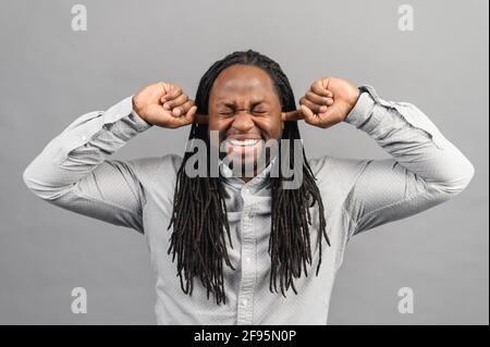 Un jeune homme afro-américain irrité, avec des dreadlocks qui bouchent les oreilles avec les doigts et qui hurle à l'œil fermé, agacé par le bruit, un homme noir ne veut rien entendre, isolé sur fond gris Banque D'Images