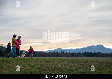 Un beau moment de famille avec des parents qui s'embrasent et des enfants qui jouent avec leur chien à l'extérieur sous le ciel du soir dans une belle prairie. Banque D'Images