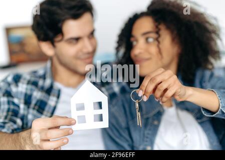 Achat d'un nouveau logement. Le couple heureux multiracial affectueux défoqué est devenu le propriétaire de leur première maison, tenant une petite maison en papier et les clés du nouvel appartement, souriant, heureux ensemble Banque D'Images