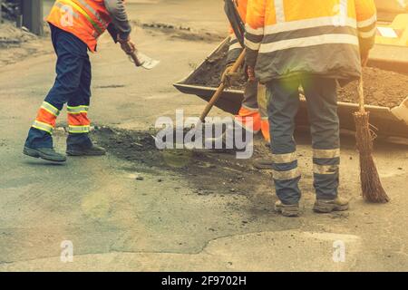 Les travailleurs de la route en uniforme réfléchissant orange vif utilisent des pelles pour gratter le sable accumulé. Entretien des chaussées et des chaussées. Réparation des nids de poule Banque D'Images