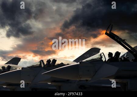 Les pilotes de avions de chasse se sont assis dans une rangée d'escadrille d'avions de guerre sur la piste coucher du soleil se préparer décollage. Spectaculaire feu rouge soleil ciel nuages silhouette avion Banque D'Images