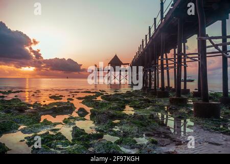 Magnifique lever du soleil et un pont en bois ou jetée menant à un restaurant / bar appelé Sansibar à la plage de Jambiani, Zanzibar, Tanzanie, Afrique à marée basse avec des roches vertes visibles dans l'eau Banque D'Images