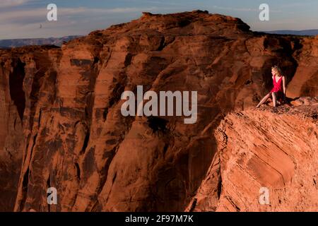 Une jeune femme blanche blonde dans une robe rouge sittung sur le bord d'une falaise à la courbe de fer à cheval, Arizona Banque D'Images