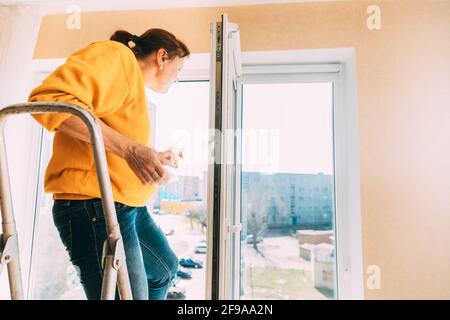 Femme de cinquante en chandail jaune et jeans lave fenêtre poussiéreuse dans l'appartement. Une femme de 50 ans nettoie les vitres des taches à l'aide de Rag and Spray Cleaner Banque D'Images