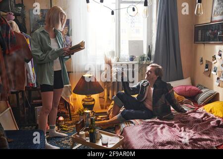 Jeune couple passant leur temps ensemble à la maison Jeune femme lire un livre et homme assis sur le lit et boire de l'alcool Banque D'Images