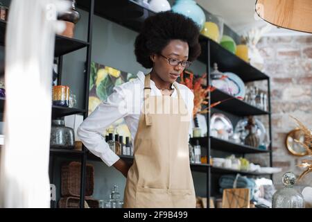 Femme afro-américaine attrayante portant un tablier beige pendant le processus de travail à la boutique de décoration. Variété de choses exclusives pour la décoration. Banque D'Images