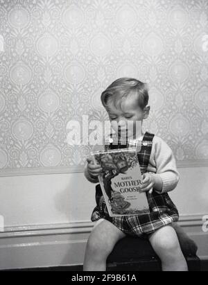 Années 1960, historique, à l'intérieur d'une chambre, un petit garçon dans une salopette tartan, assis sur un tabouret avec le livre, 'Baby's Mother Goose', Angleterre, Royaume-Uni. Banque D'Images