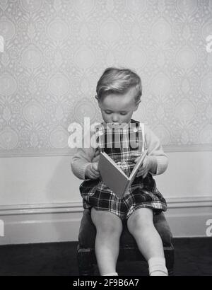 Années 1960, historique, à l'intérieur d'une pièce, un garçon de jeu de quilles portant une paire de salopettes à motif écossais, assis sur un tabouret lisant un livre, Angleterre, Royaume-Uni. Banque D'Images