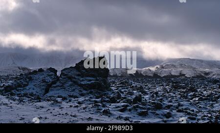 Magnifique paysage d'hiver de champ de lave couvert de neige avec des roches volcaniques et des montagnes accidentées en arrière-plan avec des nuages et la lumière du soleil en Islande. Banque D'Images