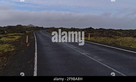 Perspective décroissante de la route sinueuse de pays avec des marques entre les champs de lave couverts de mousse de pierres volcaniques près de Grindavik, Reykjanes, Islande. Banque D'Images