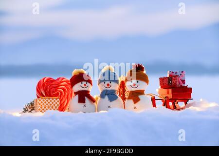 Amis bonhommes de neige restent avec un traîneau et une boîte cadeau sur la neige éclairée par des lumières de fées. Bonbons rouges à coeur blanc. Bonne Année. Composition de Noël. Banque D'Images