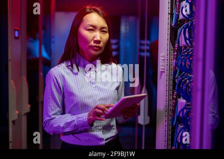 Portrait d'un ingénieur réseau asiatique sérieux qui effectue un dépannage tout en travaillant avec un superordinateur dans une salle de serveurs, lumière néon sombre Banque D'Images