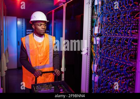 Un électricien sérieux de centre de données noir dans un casque rigide poussant le chariot avec outils lors de la réparation de l'armoire de serveur Banque D'Images