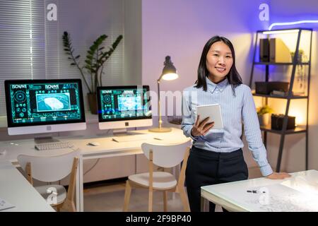 Portrait d'un beau designer asiatique souriant en chemise rayée tenir une tablette numérique dans un bureau moderne avec des ordinateurs Banque D'Images
