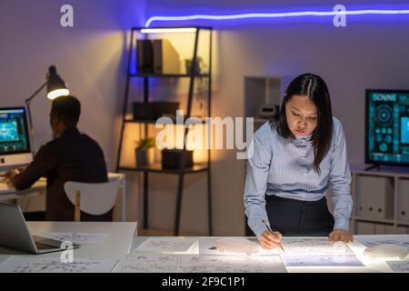 Jeune designer asiatique concentré dans une chemise habillée debout au bureau dans un bureau au néon foncé et dessin de la voiture Banque D'Images